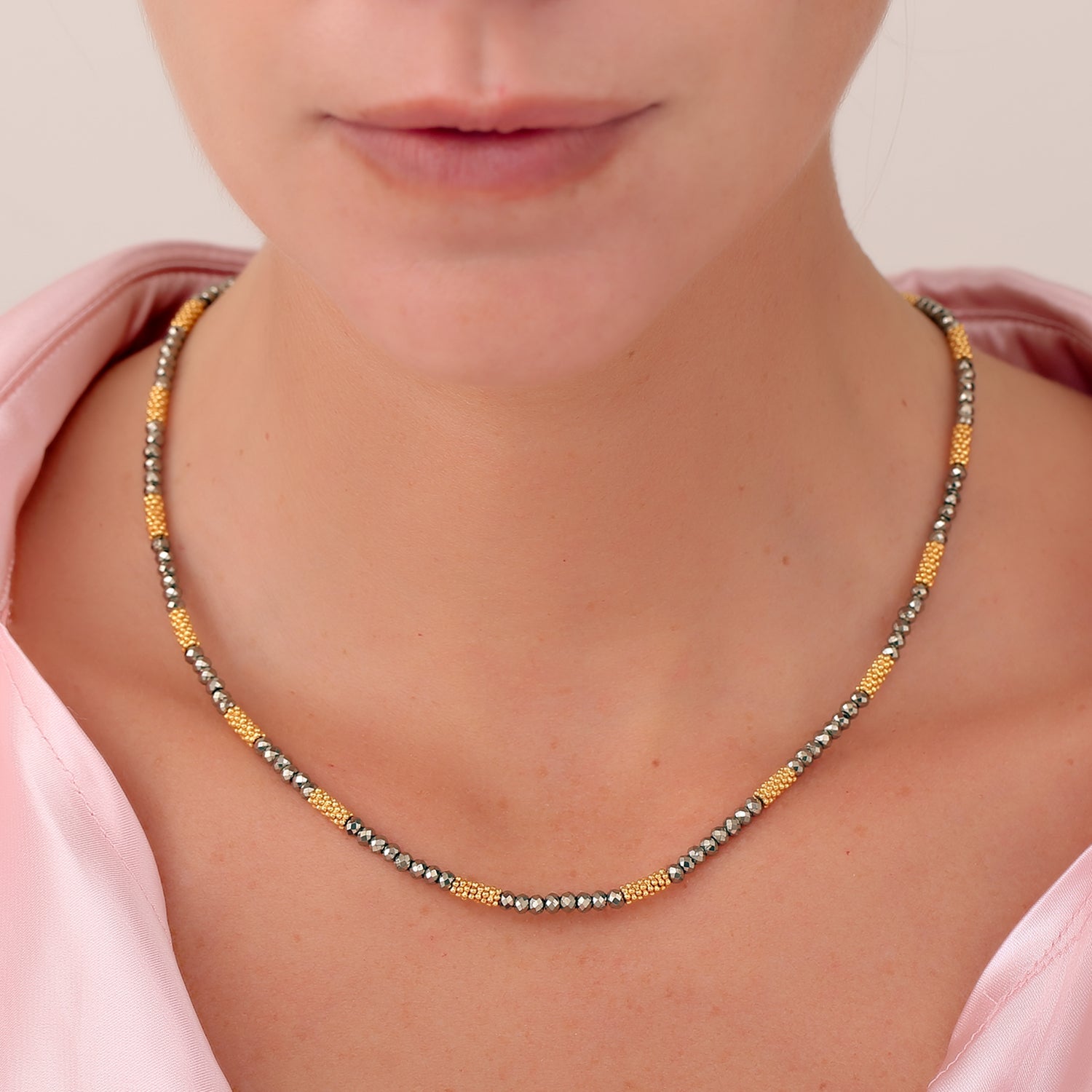 Close-up of a person wearing the Pyrite Choker, an elegant beaded necklace strung on a silk cord. It features alternating sections of small pyrites and gold-plated beads. The person is dressed in a pale pink shirt, highlighting the neckline and part of the collar against a neutral background.