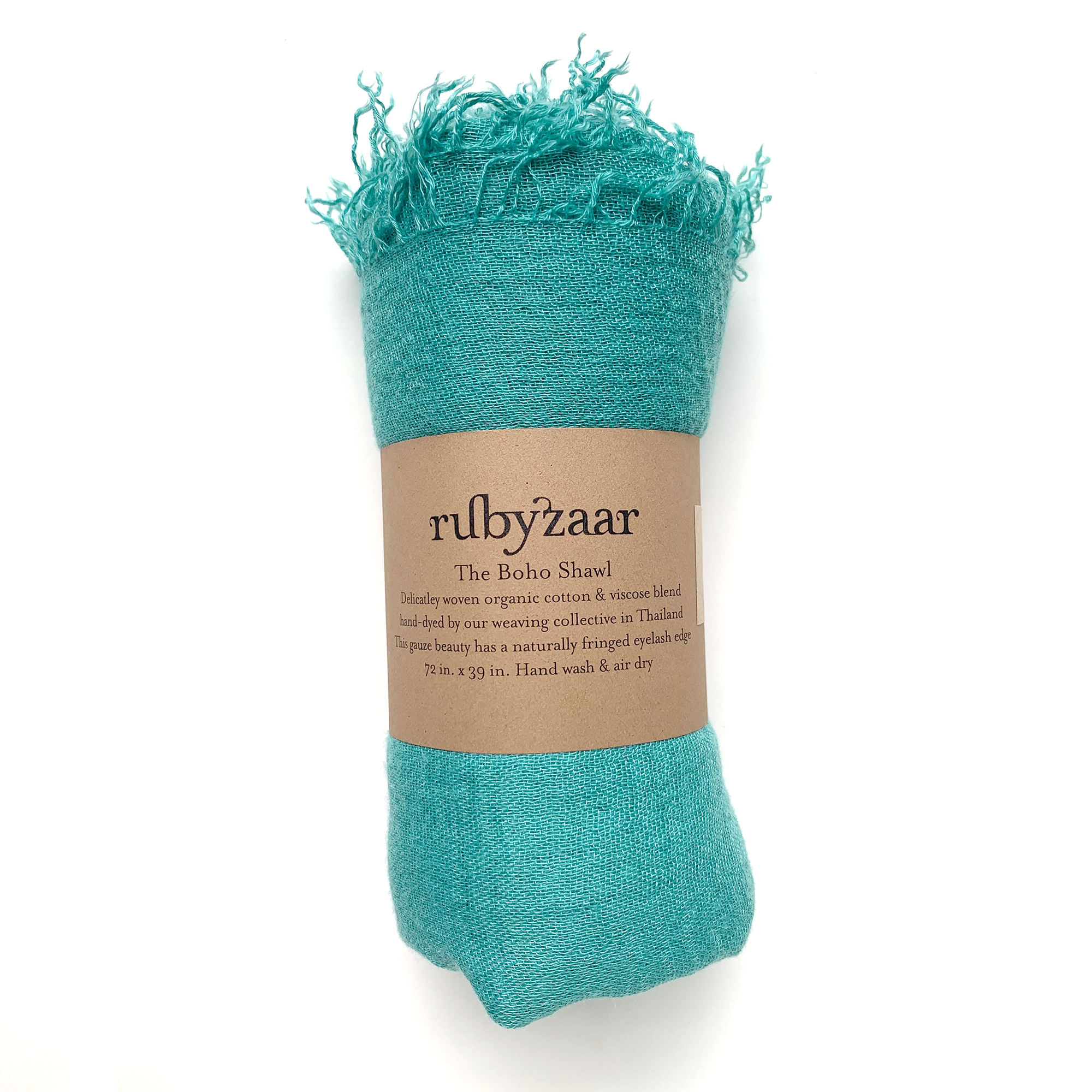 This turquoise Boho Scarf, made from a blend of cotton and viscose, showcases a bohemian style with fringed edges. It is rolled up and encased in a brown paper label that reads "rubyzzaar, The Boho Scarf," which includes details about materials and care instructions. Hand-dyed by a weaving collective in Thailand, it embodies authentic artisan craftsmanship.