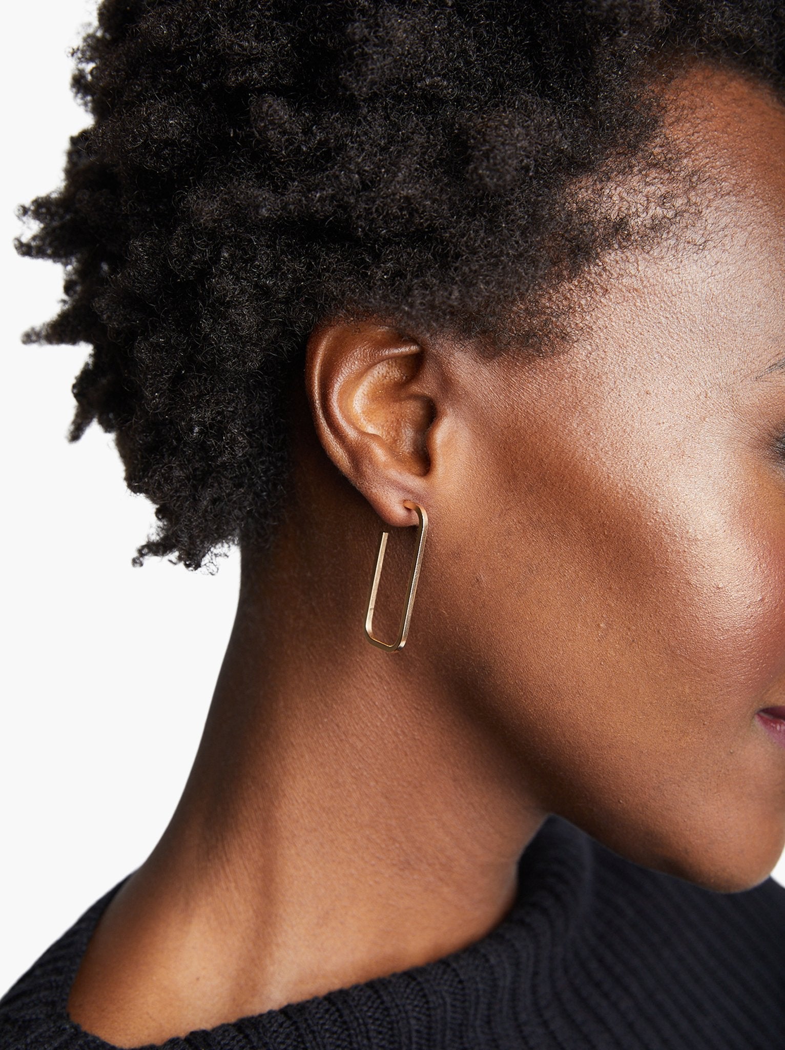Close-up of a person with short, curly hair wearing a single rectangular Bali Hoop earring crafted from 14k gold-fill. Their dark skin tone complements the black textured top, while the white, uncluttered background highlights the elegance of this handmade jewelry piece.