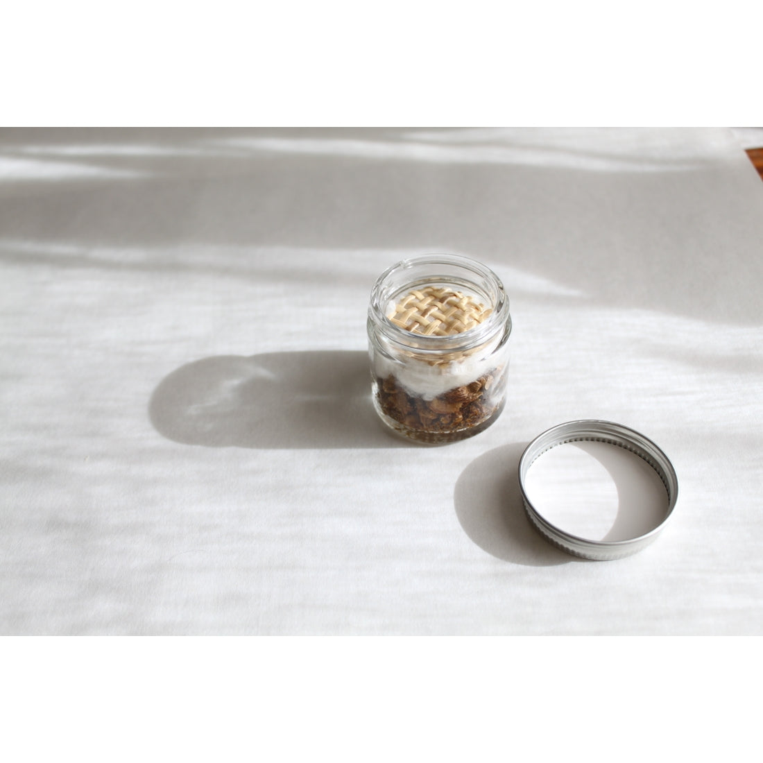 An Aromatherapy Inhaler from the collection of 13 varieties is open on a white surface. Inside, a blend of dried herbs suggests an aromatic stress relief aid. The lid rests nearby, casting light and shadow patterns that add texture to the scene, reminiscent of a calming essence in the air.