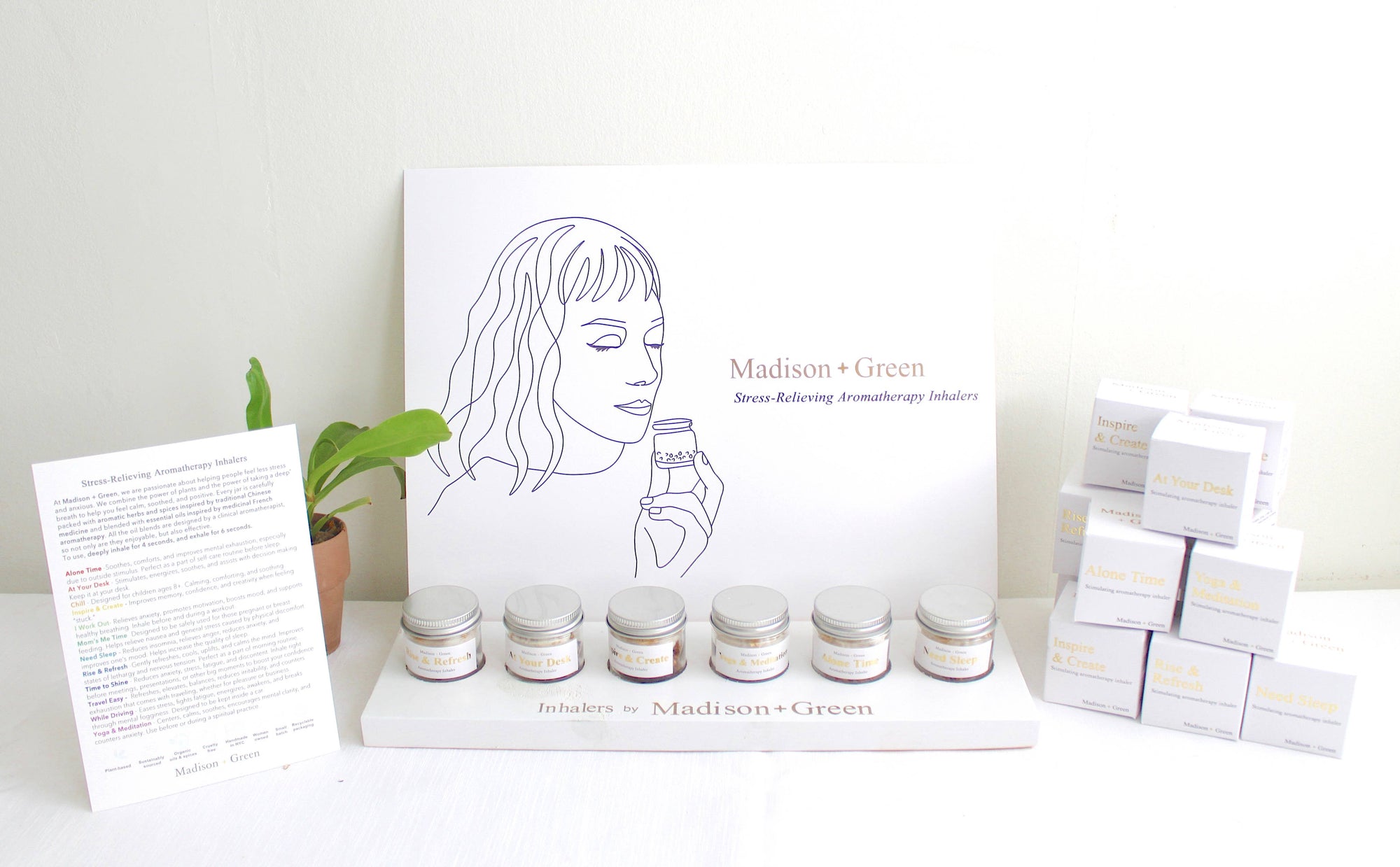 A display of Aromatherapy Inhalers featuring 13 varieties with stress-relieving organic essential oils. Six jars are neatly arranged on a stand in front of an illustrated sign. On the left, there is a potted plant and informational pamphlet, while stacked product boxes are positioned on the right, all set against a simple, light-colored wall.