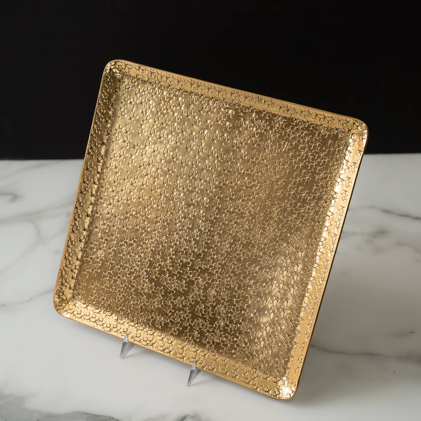 The Gold Aluminum Hammered Square Tray - 11” is crafted from sustainable materials and showcases an intricate textured surface with delicate star-like patterns. Its raised edge stands out elegantly on a white marble surface against a black backdrop, and its reflective shimmer adds a touch of sophistication to any decor.