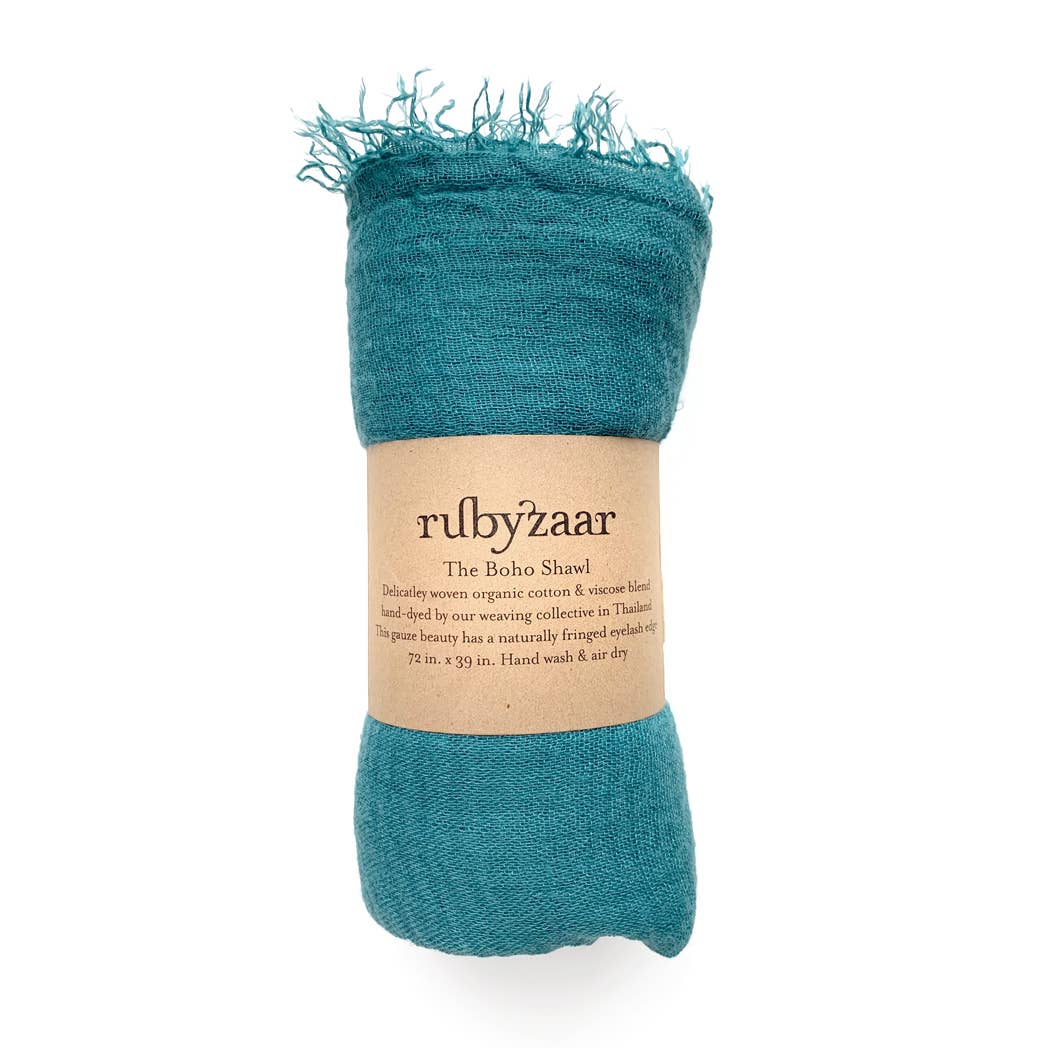 A teal blue scarf, capturing a bohemian essence, is neatly rolled with a beige label that says "rubyzzaar" and describes it as "The Boho Scarf." This hand-dyed cotton viscose scarf measures 72 by 29 inches, featuring fringed edges and care instructions for hand washing and air drying.