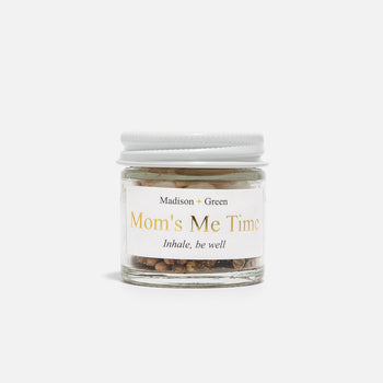 A small clear jar with a white lid, filled with stress-relieving herbs and organic essential oils, is labeled with "Madison + Green" at the top and "Mom's Me Time" in gold text, followed by the phrase "Inhale, be well." The Aromatherapy Inhaler is showcased against a plain white background.