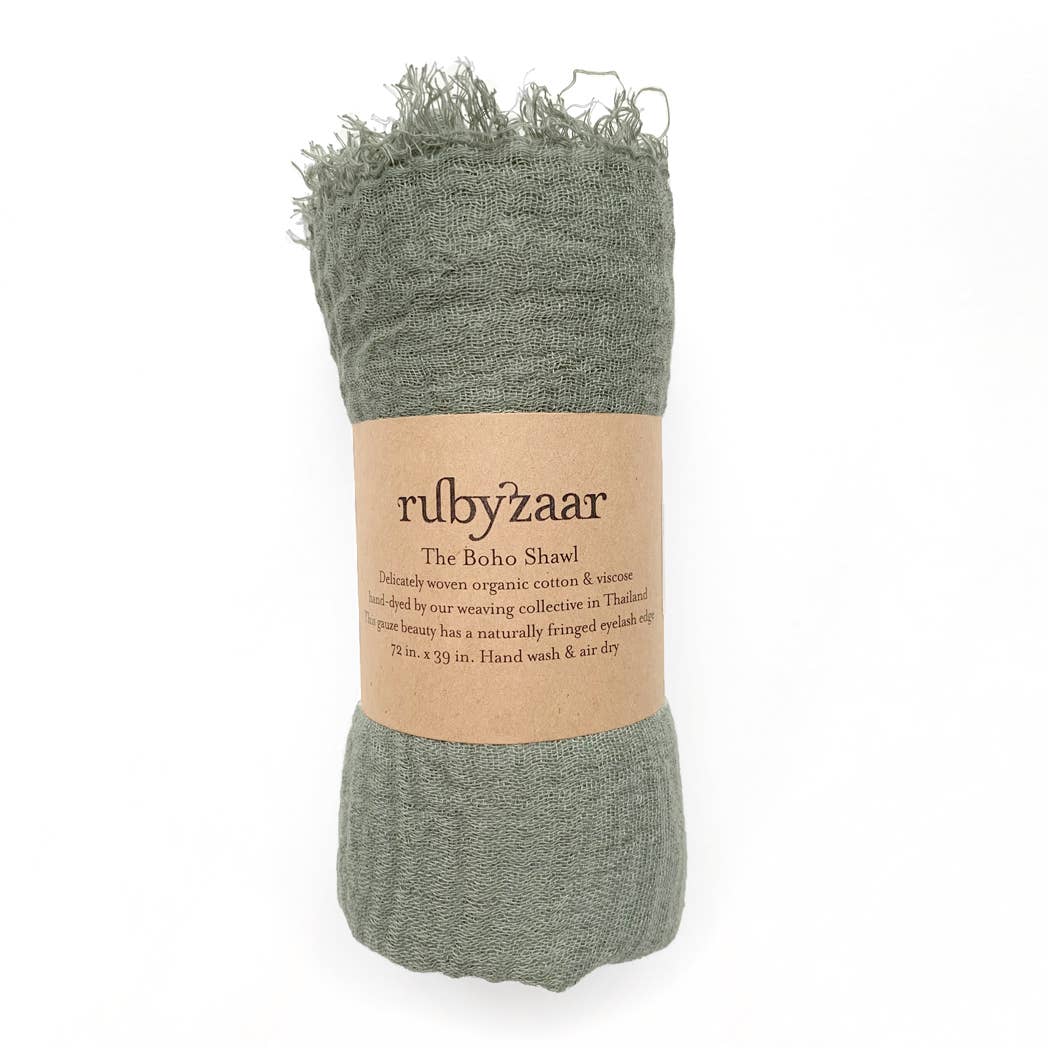 A rolled-up green scarf with a natural fringe is wrapped in a brown paper label. The label reads "rubyzaar," "The Boho Scarf," and describes the hand-dyed, bohemian style item as delicately woven from organic cotton and viscose. It measures 72 x 39 inches, is hand wash only, and made in Thailand.