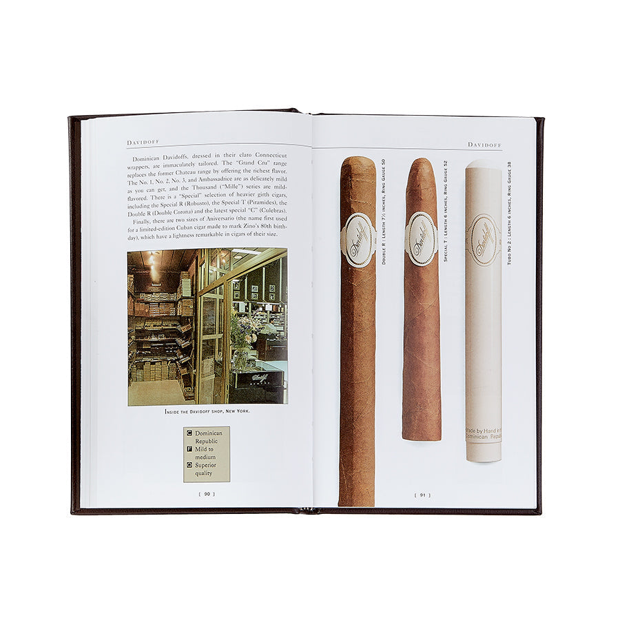 An open book reveals the wisdom of The Cigar Companion (Brown Bonded Leather), showcasing an enticing image of a cigar store interior. On the opposite page, three handcrafted cigars—a dark, medium, and light—are presented against a pristine white backdrop. Each cigar is clearly labeled, offering a genuine pleasure for any enthusiast or reader of The Cigar Companion.