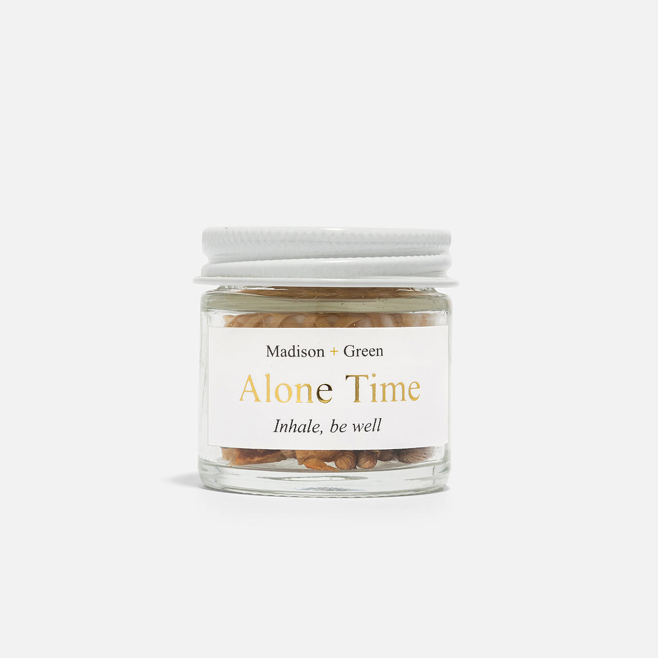 A small glass jar with a white lid, filled with dried botanical ingredients and essential oils, is featured. The label displays "Madison + Green" and "Alone Time," followed by the phrase "Inhale, be well." This particular inhaler from the Aromatherapy Inhalers' 13 varieties collection serves as an effective stress reliever, inviting a moment of calm against a plain white backdrop.