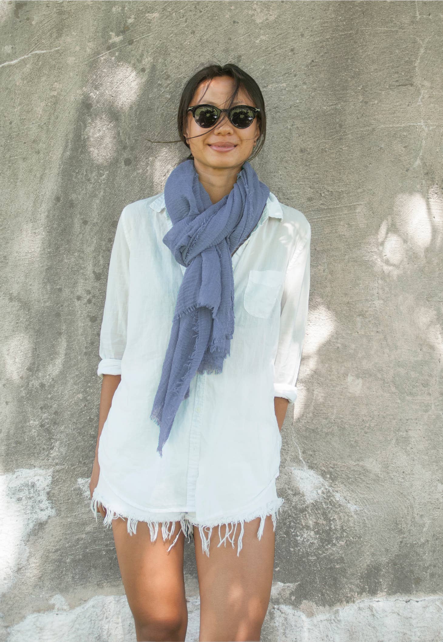 A person wearing sunglasses smiles while standing against a textured stone wall. They are dressed in a light white button-up shirt with the sleeves rolled up, white fringed shorts, and a large Boho Scarf in hand-dyed bohemian style. Sunlight creates dappled shadows on the wall and ground.