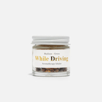 A small glass jar from the Aromatherapy Inhalers collection by Madison + Green, featuring stress-relieving scents, contains visible herbs or seeds and is topped with a white screw cap. The label showcases minimalistic black and gold lettering on a white background, set against a plain white backdrop.