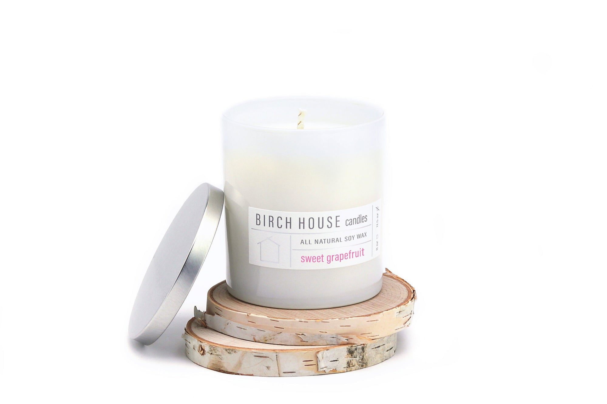 A "Sweet Grapefruit Candle" from Birch House Candles features a citrus blend of premium fragrance oils in frosted glass, nestled on birch wood slices. A silver lid leans nearby, and the plain white background highlights its minimalist design.