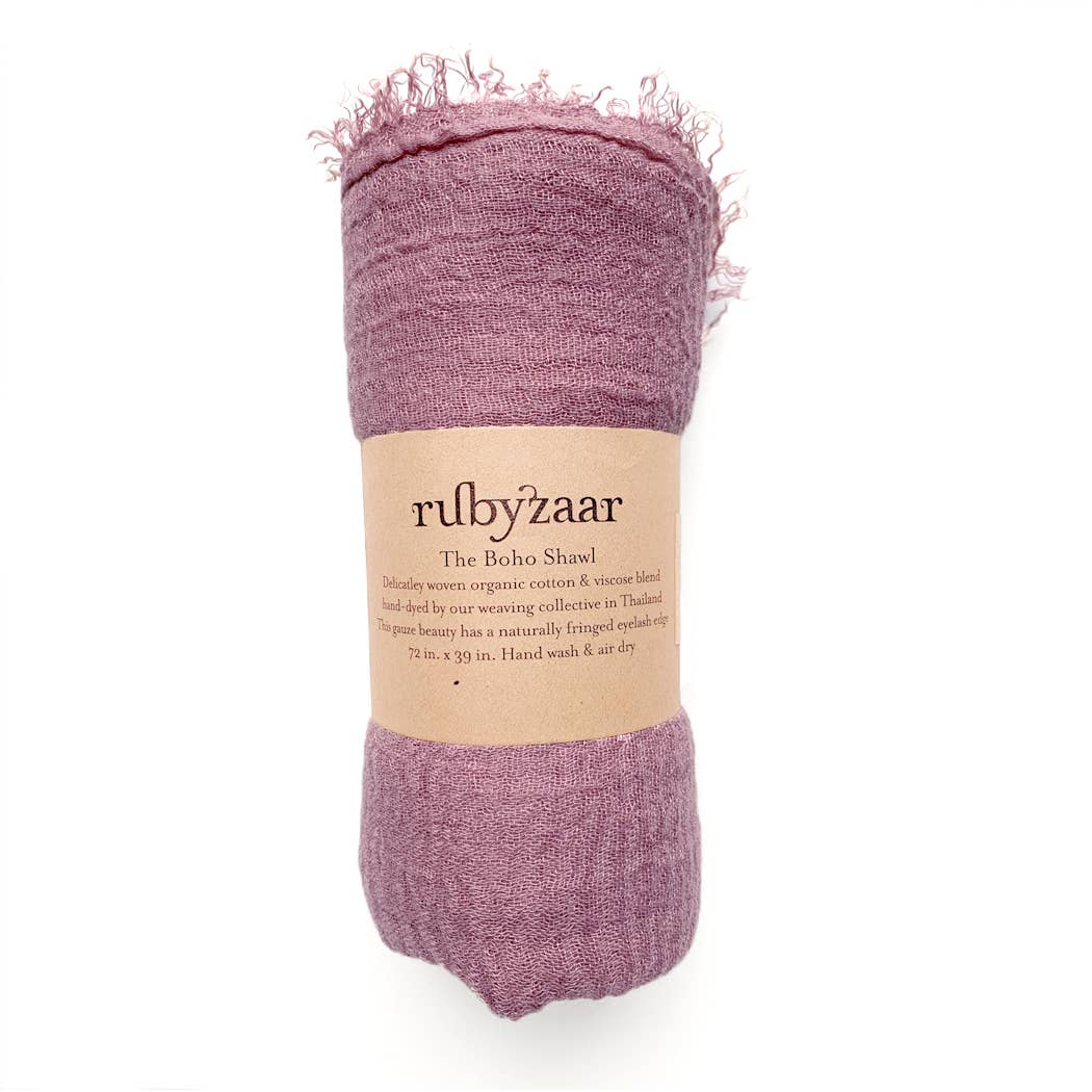 A rolled-up pink hand-dyed Boho Scarf from rubyzaar, fashioned from organic cotton and viscose. It bears a tag celebrating its handmade craftsmanship by a weaving collective in Thailand. This bohemian-style shawl showcases naturally fringed edges and measures 72 inches by 39 inches. Hand washing and air drying are recommended for care.