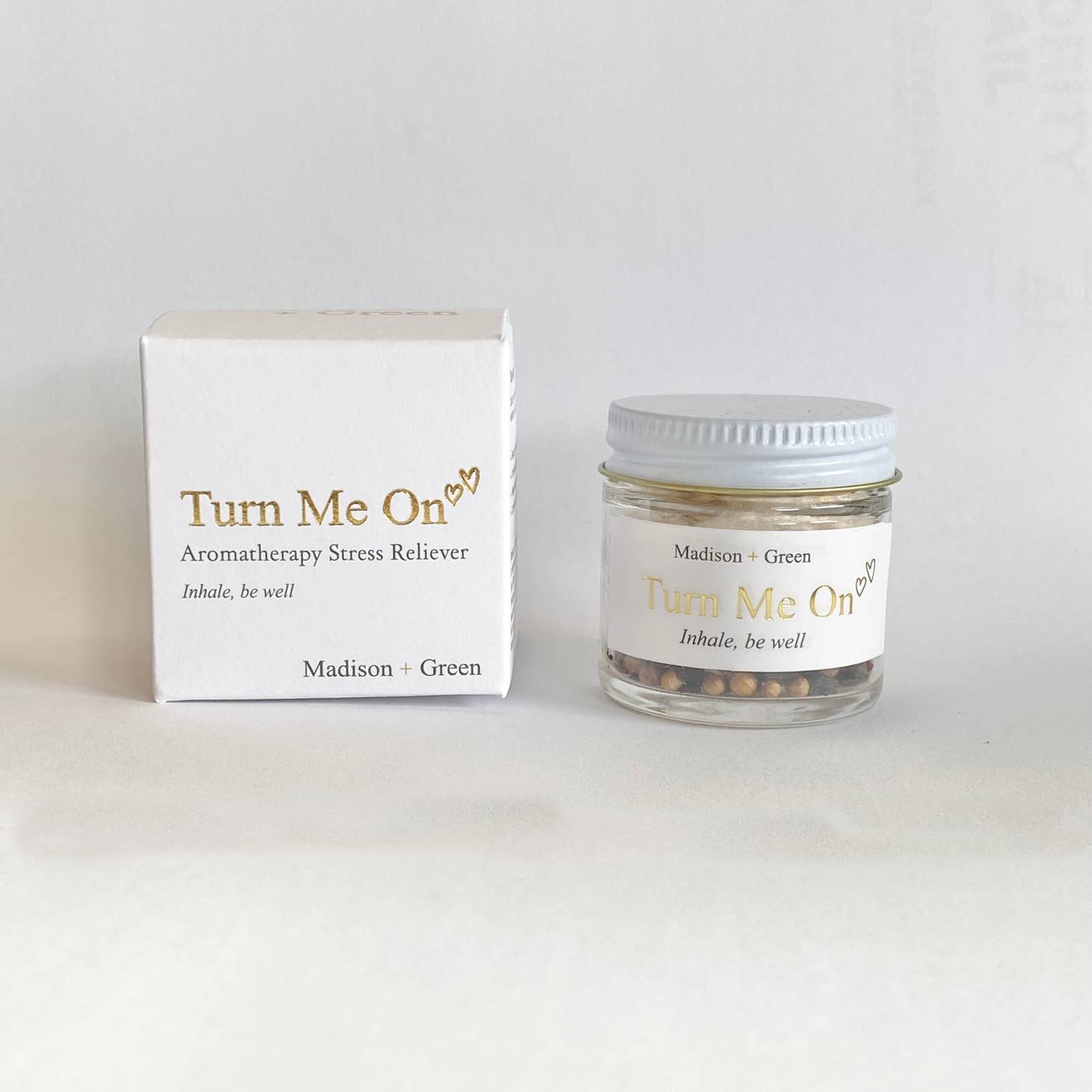 A small glass jar with a white lid, labeled "Turn Me On," is placed beside a matching white box. Both are adorned with gold text reading "Aromatherapy Stress Reliever. Inhale, be well," from "Madison + Green." The jar contains aromatic beads infused with organic essential oils designed for stress-relieving scents. This product is part of Madison + Green's Aromatherapy Inhalers collection, featuring 13 different varieties.