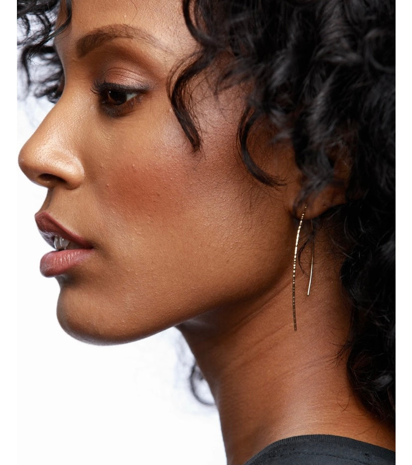 A close-up profile of a person with curly hair highlights the minimalist Galaxy Earrings, crafted from 14k gold-fill. Their sleek, elongated design accentuates the sharpness of their features and embodies ethical fashion. The earrings stand out beautifully against their hair, creating an empowering visual contrast.