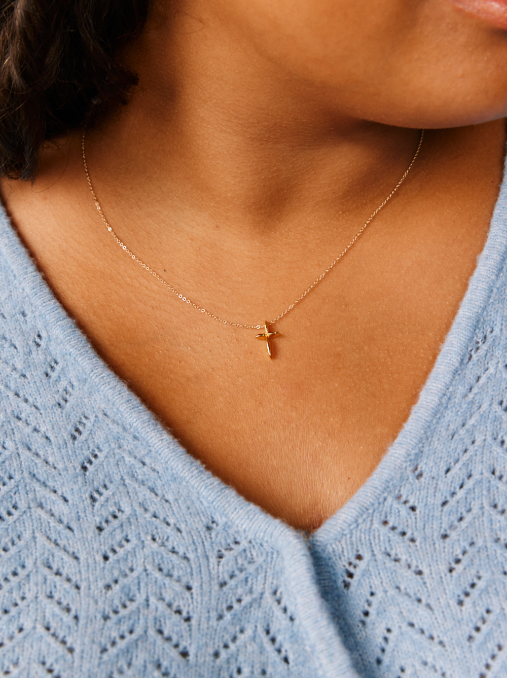 A close-up showcases a person adorned with the Droplet Cross Necklace, a delicate vermeil piece featuring a handmade pendant crafted in Nashville. The individual wears a light blue V-neck knit sweater that complements and enhances the elegance of the necklace. The focus remains on its intricate design, while the textured sweater adds subtle depth to the image.