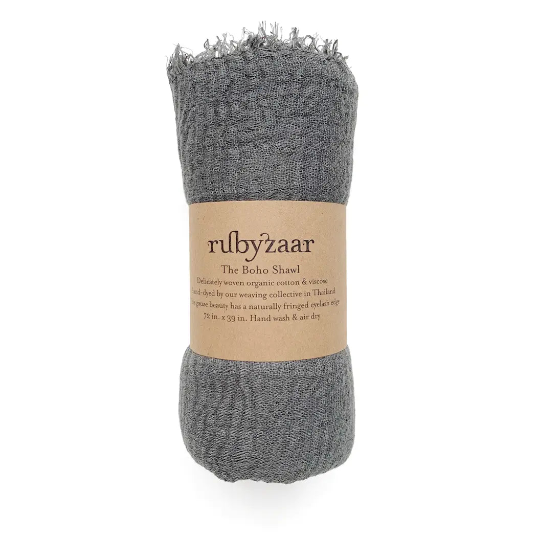 A gray Boho Scarf with a fringed edge is rolled up and wrapped in a brown kraft paper label. The label bears the name "rubyzaar" and describes it as being made from hand-dyed organic cotton and viscose. It emphasizes its artisan craftsmanship, along with care instructions: "Hand wash & air dry.