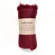 A deep red Boho Scarf with frayed edges is elegantly rolled up and wrapped in a beige label. The label reads "rubyzaar" and details that the scarf, designed by Wendy Pernu, is handwoven in Northern Thailand from 100% textured cotton.