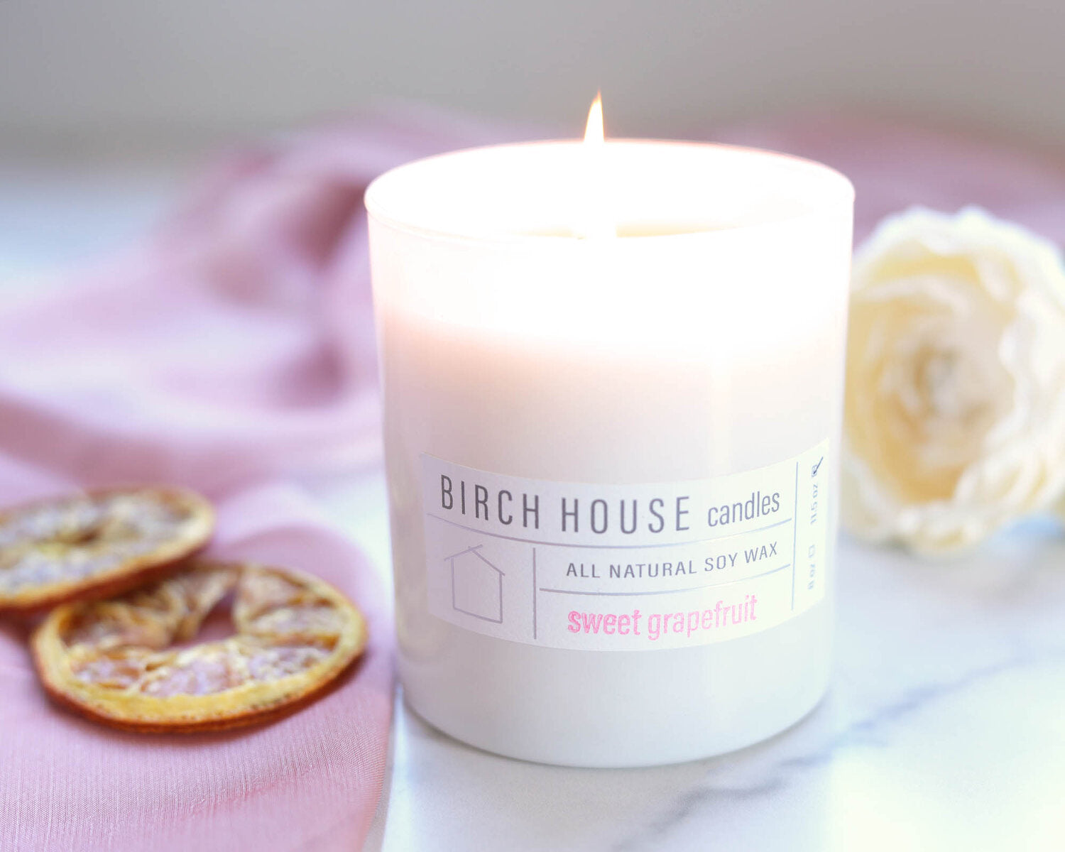 A "Sweet Grapefruit" soy wax candle by Birch House Candles, in a glass jar, sits on soft pink cloth with dried grapefruit slices. A blurred white rose graces the backdrop on a smooth white surface, exuding premium fragrance oils.