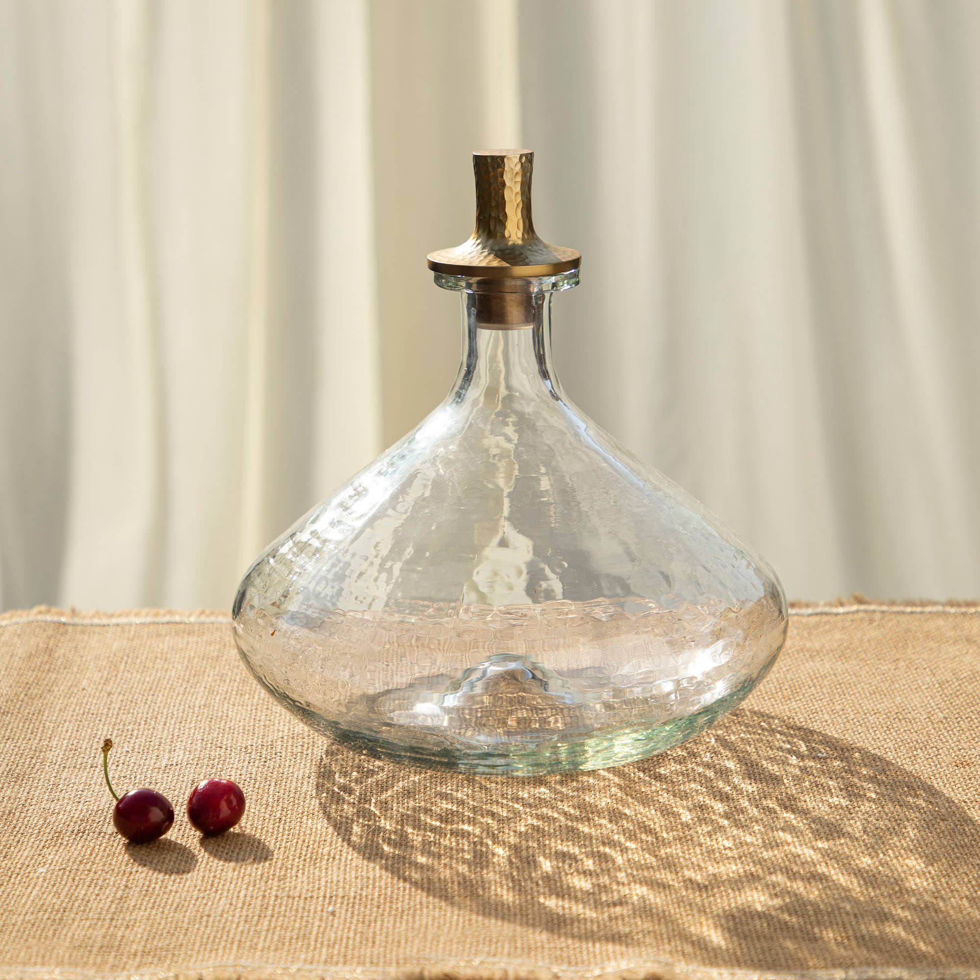 The Decanter Wide, featuring a clear pebbled glass design and a gold-finished stopper, rests on a textured beige surface, casting intricate shadows. Nearby, two cherries add to the scene while softly blurred light-colored drapes in the background create a warm, elegant ambiance.