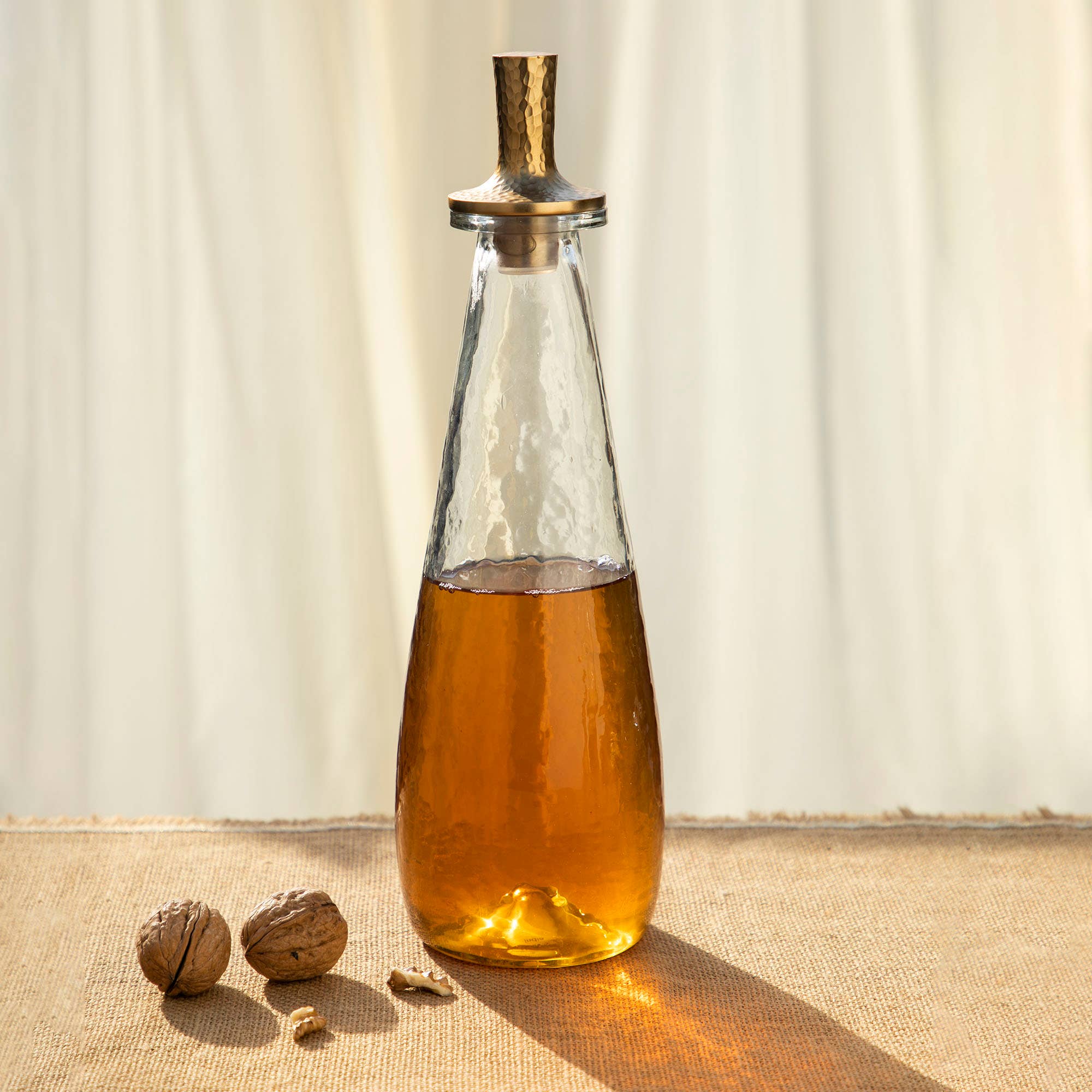 A Decanter Tall, crafted from clear glass and filled with an amber liquid, is topped with a textured gold cap and rests on a beige cloth. Nearby, a pair of whole walnuts and some walnut pieces are spread around. The background offers soft, diffused light filtering through a sheer curtain, enhancing the warm and inviting ambiance of the dining area.