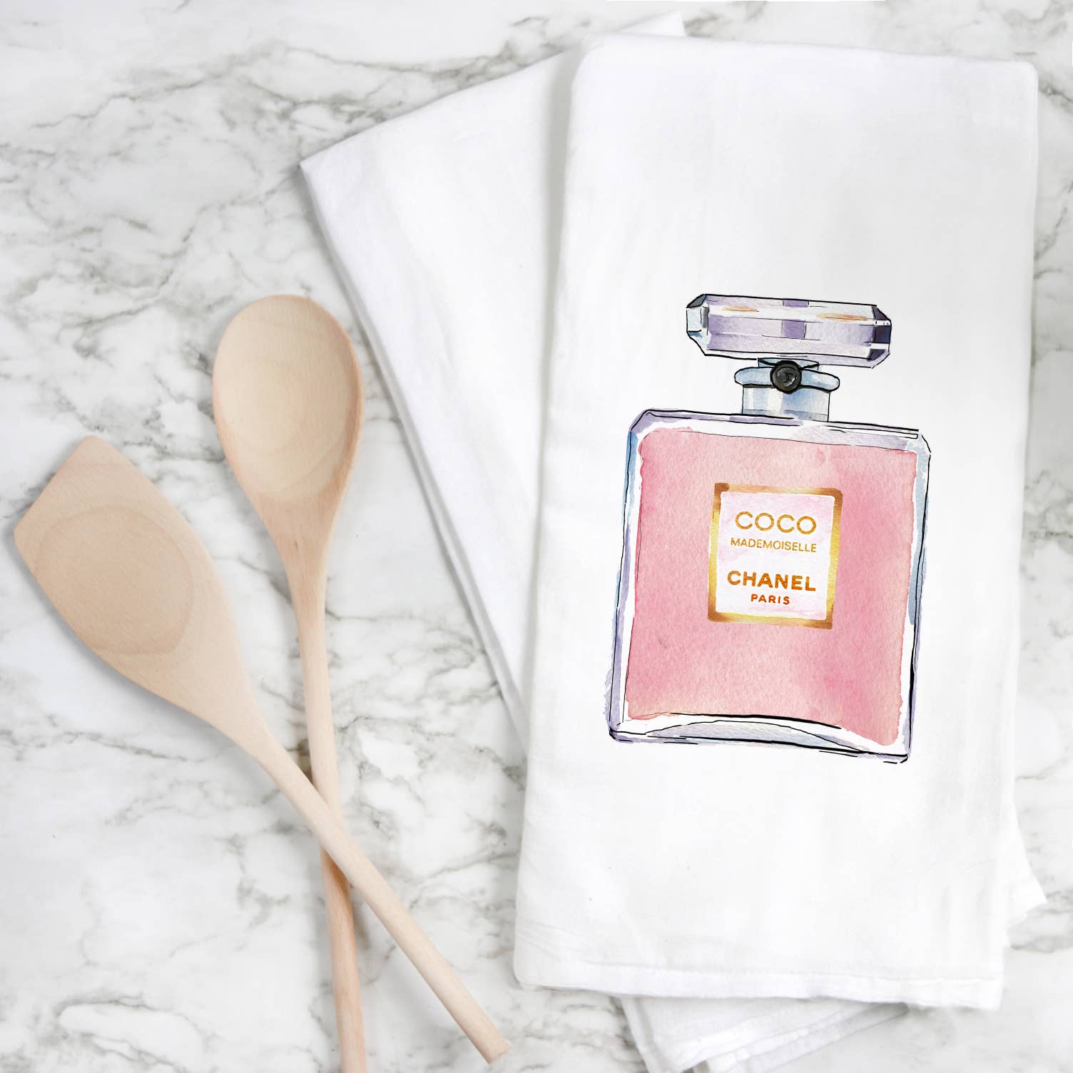 A marble surface showcases two elegant white kitchen towels from the "Tea Towels - Chanel" collection, uniquely designed with an illustrated pink bottle of Chanel Coco Mademoiselle perfume. Alongside them are two handcrafted wooden cooking spoons, emphasizing both elegance and culinary simplicity in the composition.
