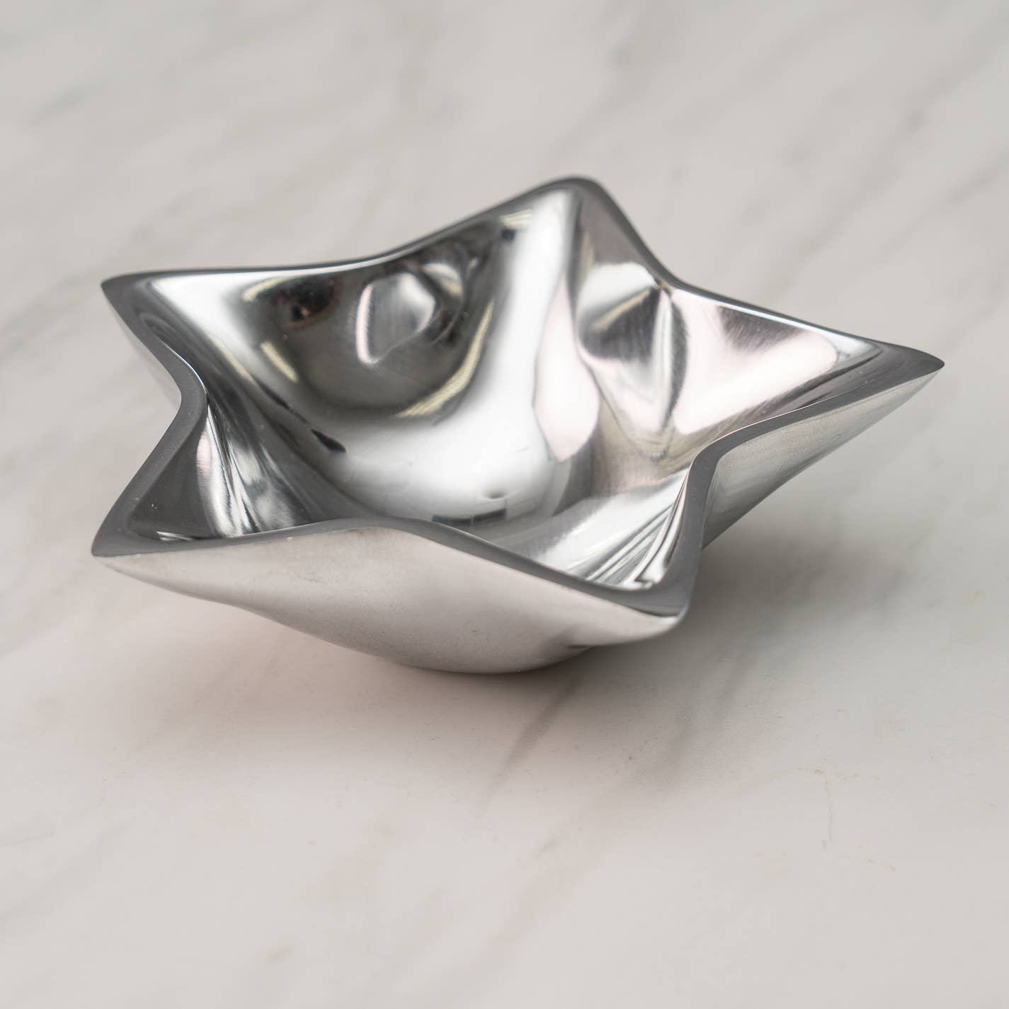 The Aluminum Star Tray, crafted with smooth and curved edges, features a reflective silver aluminum finish that rests elegantly on a white marble surface. Its shiny metallic appearance underscores its modern and sleek design, making it food safe yet best maintained by hand washing to preserve its dazzling look.