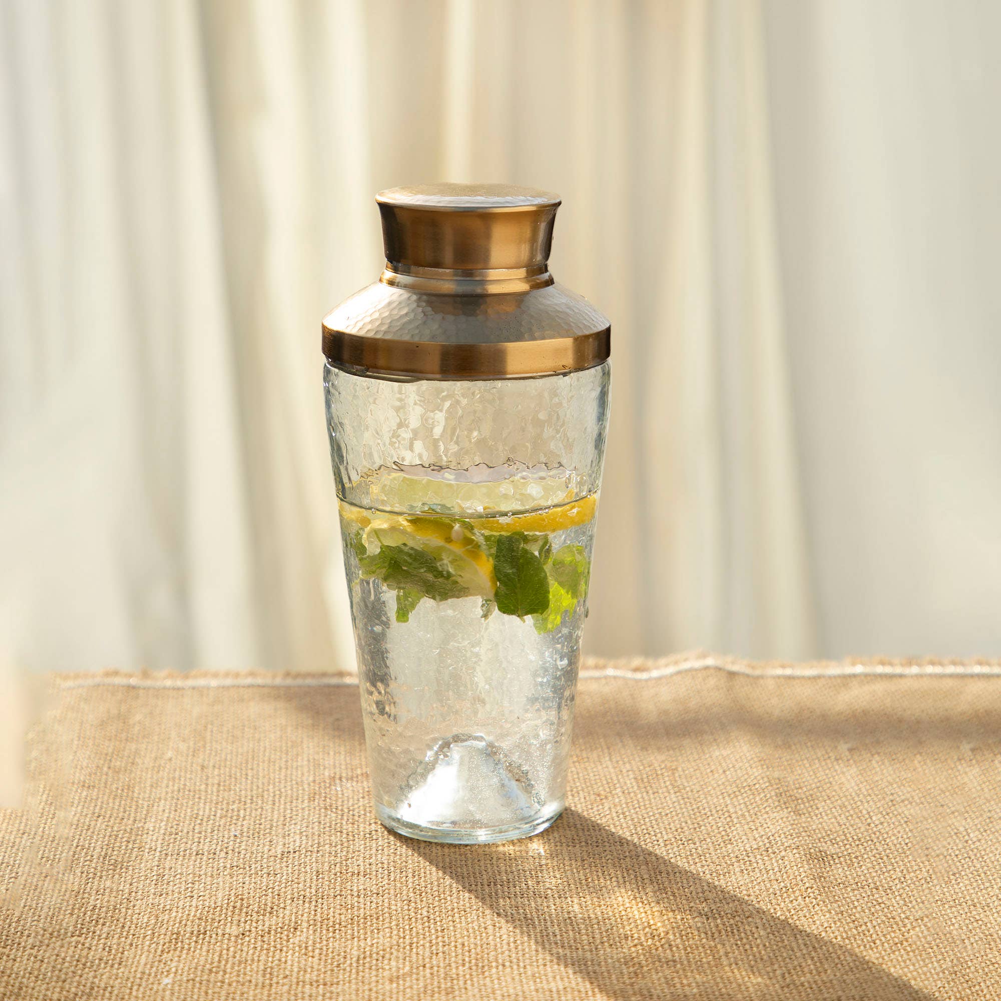 A Cocktail Shaker with a golden metal lid sits on a textured burlap surface. Inside, slices of lime and mint leaves float in clear water. The softly blurred beige and white background creates a warm, inviting atmosphere, perfect as part of your home bar essentials.