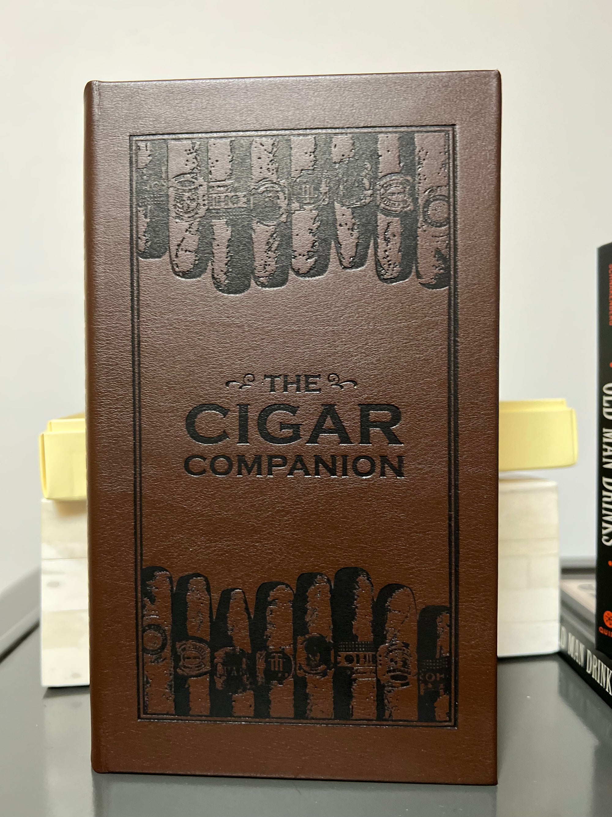 The image showcases "The Cigar Companion" in brown bonded leather, a must-have for cigar enthusiasts, adorned with a pattern of handmade cigars at the top and bottom. It is positioned upright on a gray surface, with other books partly visible in the background.