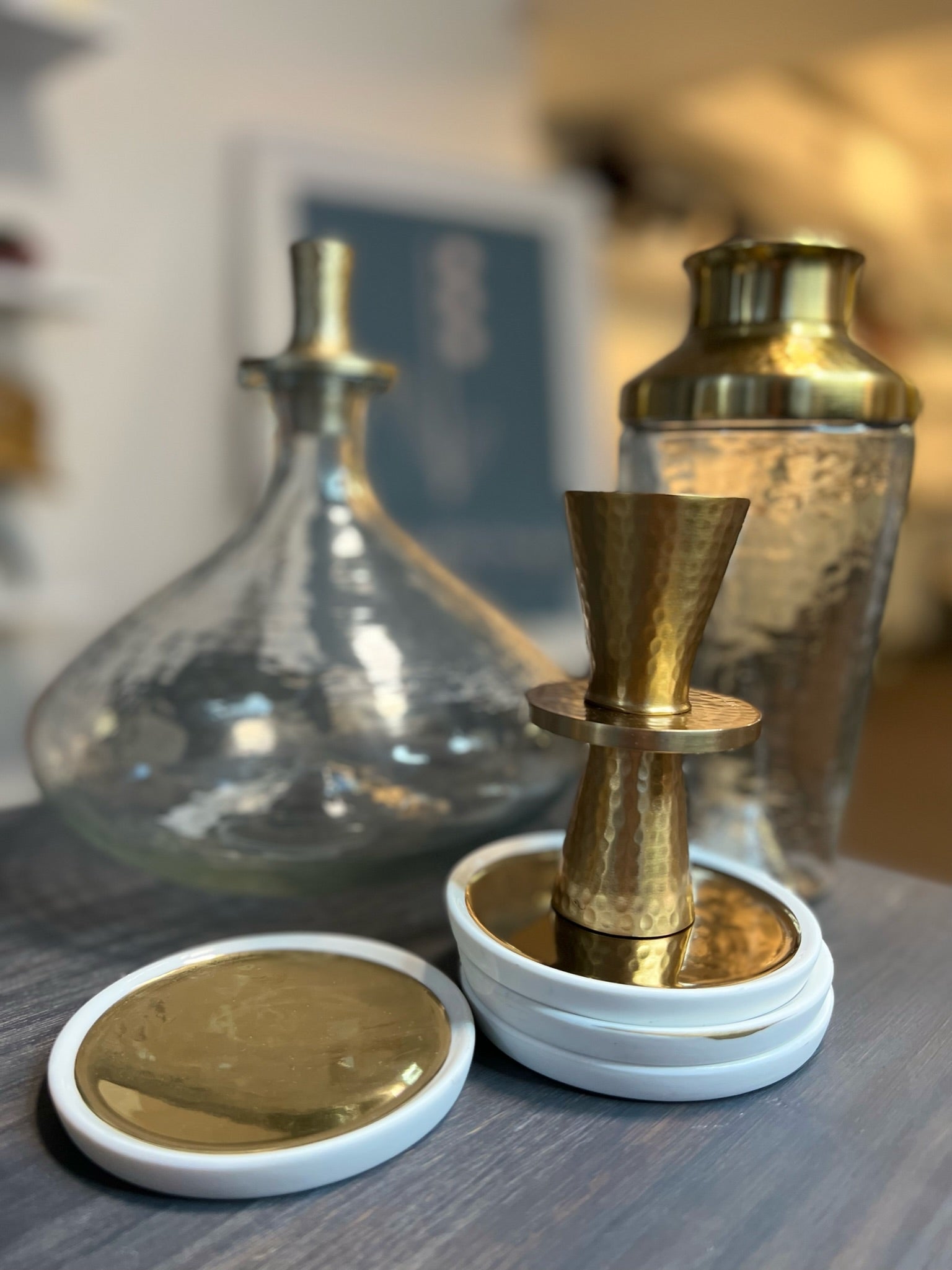 A collection of elegant barware on a wooden surface includes the Decanter Wide, featuring a wide pebbled glass design with an exquisite gold stopper, alongside a hammered metal cocktail shaker and a gold double jigger. The scene is completed by several white coasters with gold tops, all set against a softly blurred background that highlights their textures.