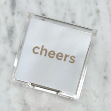 Load image into Gallery viewer, A white square coaster featuring the word &quot;cheers&quot; in gold lettering rests on a light gray marble surface, making it an ideal hostess gift. The coaster is part of the &quot;Cocktail Napkin Acrylic Tray Set - Year Round Entertaining&quot; and is encased in a transparent acrylic holder.
