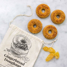 Load image into Gallery viewer, Four gingerbread doughnuts, made with the Gingerbread Doughnut Baking Mix &amp; Mold Set, are sprinkled with crumb topping and elegantly arranged on a marble surface. Next to them is a beige drawstring bag featuring a doughnut illustration and the words &quot;Gingerbread Doughnut.&quot; A few pieces of candied ginger finish off the scene.
