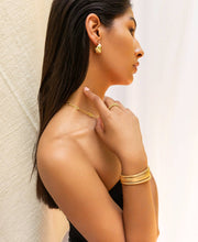 Load image into Gallery viewer, A woman poses against a light background, elegantly displaying her jewelry. She wears gold hoop earrings with a 3 mm hinge closure, a chain necklace, and the Flat Hinge Bangle on her wrist. Her long, dark hair cascades down her back as she touches her neck to accentuate her ring and bangle. She is dressed in a black strapless top.
