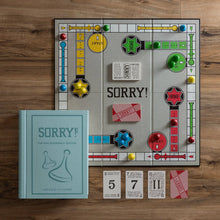 Load image into Gallery viewer, The Sorry! Vintage Bookshelf Edition is displayed on a wooden table, featuring vibrant paths, a &quot;Home&quot; zone, and four starting areas. Red, blue, green, and yellow pawns are ready for play. The game includes number and &quot;Sorry!&quot; cards and comes in an elegant fabric-wrapped book-style box—ideal for any collectible bookshelf.
