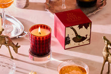 Load image into Gallery viewer, A red-patterned Poinsettia Candle burns on a reflective holder. Nearby, a red box labeled &quot;Poinsettia&quot; with a turkey design sits on the table. A silver tray hosts drinks such as Poinsettia cocktails in glasses alongside brass animal figurines. The warmly lit scene creates a cozy, festive atmosphere.
