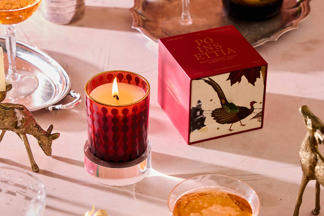 A red-patterned Poinsettia Candle burns on a reflective holder. Nearby, a red box labeled 