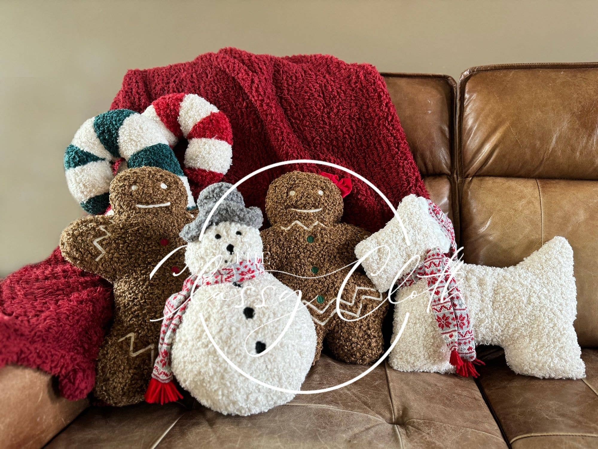 A cozy holiday-themed setup on a brown leather couch features plush decorations: two gingerbread figures, a snowman, and a candy cane—all textured and fluffy. The Christmas Sherpa Pillow Gingerbread Man adds charm beside the red knit blanket, which drapes over the couch, enhancing the festive warmth with an inviting touch.
