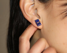 Load image into Gallery viewer, A close-up showcases an ear wearing a pair of Lapis Studs, handcrafted with dark blue stones. The earring design includes two stacked square lapis stones separated by a 14k gold-filled band. A hand gently touches the earlobe, set against a neutral background that accentuates the studs&#39; intricate details.
