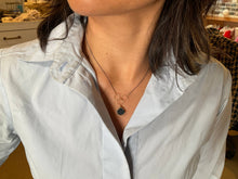 Load image into Gallery viewer, A person in a light blue collared shirt models the Cassiopeia Necklace, which is an adjustable piece featuring a black pendant and three interconnected gold rings. The blurred backdrop of household items and furniture accentuates the necklace&#39;s elegant design.
