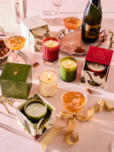 Load image into Gallery viewer, A festive tabletop scene features lit Poinsettia Candles in decorative holders, champagne glasses, and a poinsettia cocktail on a tray beside the sparkling wine. A gold ribbon elegantly sprawls across the table amid candle boxes, creating a warm and celebratory atmosphere.
