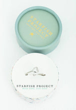 Load image into Gallery viewer, The Ada Silver Heart Ring, with its adjustable design for a perfect fit, is elegantly showcased atop a round box. The lid of the box, in light teal, is adorned with &quot;Starfish Project&quot; in gold lettering. This text is echoed on both the top and bottom sections against a white, textured surface.
