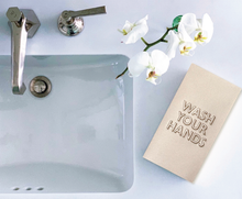 Load image into Gallery viewer, A bathroom sink with a chrome faucet and soap dispenser is shown. To the right, there is a blooming white orchid. Next to it is a beige Cloth-Like Guest Towel featuring the &quot;WASH YOUR HANDS&quot; embroidery in large capital letters.
