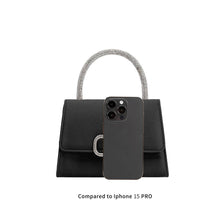 Load image into Gallery viewer, The Sadie Satin Top Handle Bag, a small black handbag with a rhinestone top handle and rectangular flap, is displayed alongside a black iPhone 15 Pro for size comparison. The silver buckle detail on the flap adds elegance. Text below reads &quot;Compared to iPhone 15 PRO.
