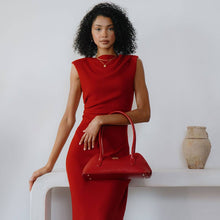 Load image into Gallery viewer, A curly-haired woman stands against a white background wearing a sleeveless red dress, holding a Dakota Recycled Vegan Shoulder Bag. Behind her, a simple white shelf holds a beige ceramic vase. The minimal and elegant setting is enhanced by the bag&#39;s OEKO-TEX certified lining.
