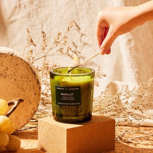 A hand lights a Merlot Candle 6 oz on a cork platform with yellow grapes and dried grass, evoking a rustic wine lover's scene against a beige fabric backdrop with a round stone slab.