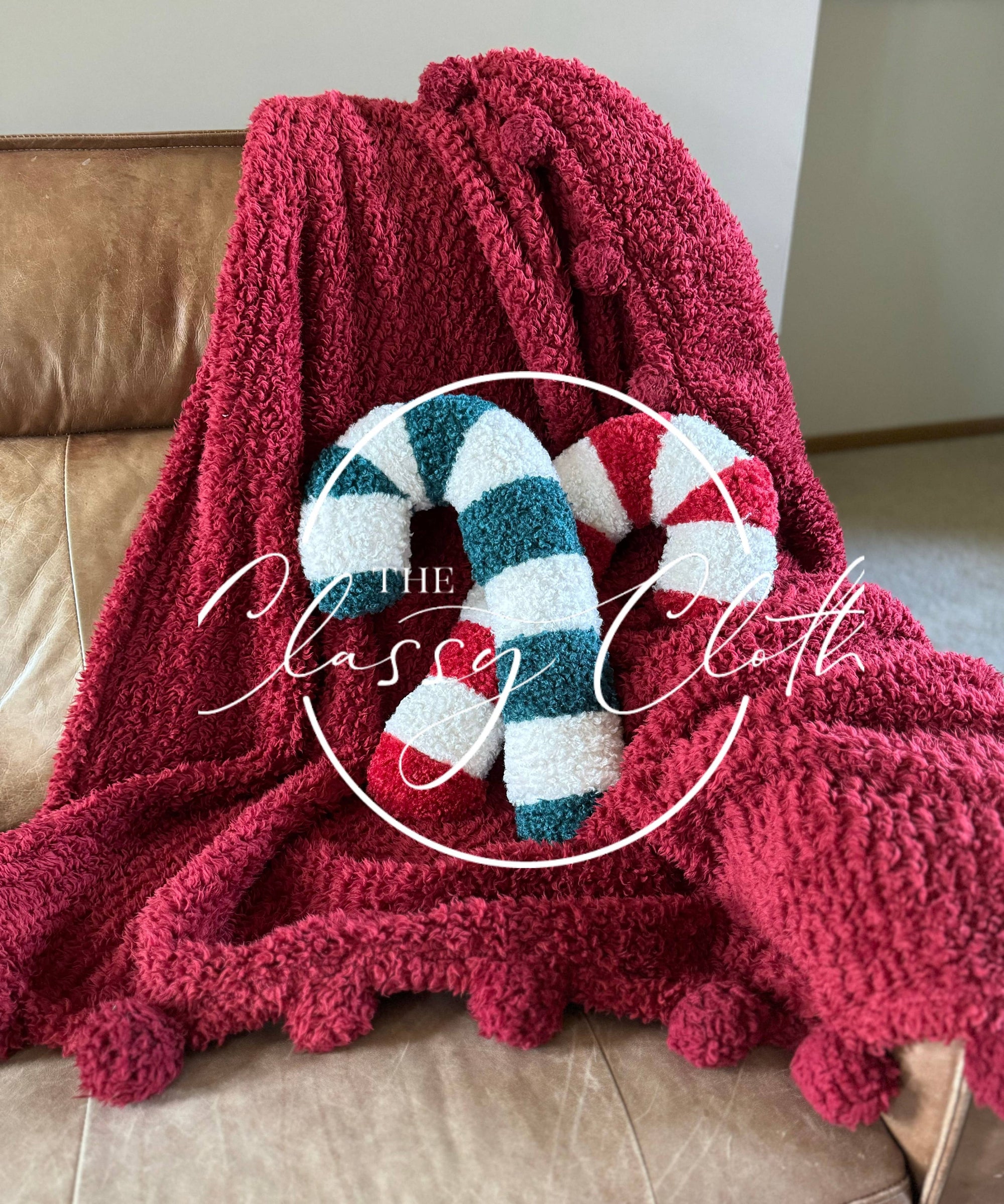 Draped over a beige leather chair is a plush red blanket adorned with pom-pom edges, accompanied by the Christmas Sherpa Pillow Red Candy Cane. The words "The Classy Cloth" elegantly overlay the image in cursive within a circle, making it perfect for your holiday decorations.