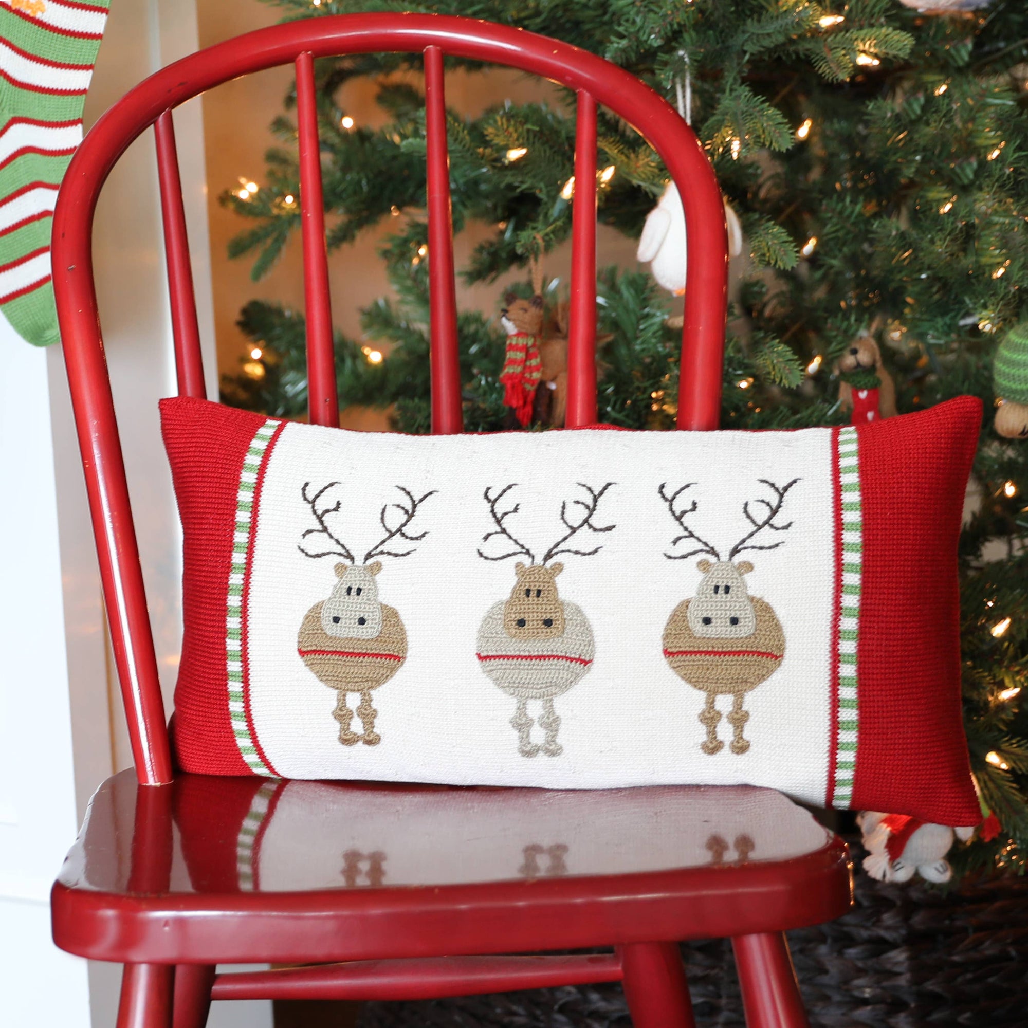 A red wooden chair is adorned with a festive Round Reindeer Lumbar Pillow, which features three cartoon reindeer wearing striped scarves on a white background and boasts green and red striped borders reminiscent of traditional Armenian crafts. In the backdrop, a partially visible Christmas tree decorated with lights and ornaments adds to the holiday spirit, accompanied by a green stocking hanging nearby.