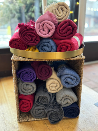 A woven basket on a wooden floor holds Boho Scarves in vibrant colors like red, pink, and purple. These hand-dyed cotton & viscose treasures are stacked both in the basket and on top. A window beyond reveals a blurred outdoor scene.