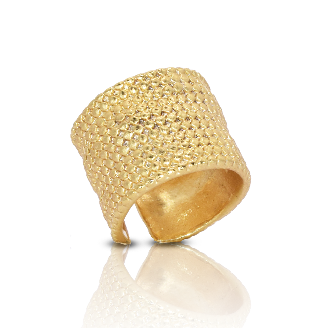 The Chainmail Ring is a wide piece with 24 kt gold plating and a textured, hammered surface that reflects light to create a shimmering effect. Slightly tilted on a smooth surface, it enhances its golden appearance and makes a bold statement.