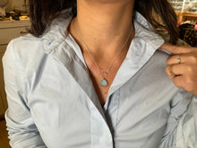 Load image into Gallery viewer, Close-up of a person in a pale blue collared shirt, partially unbuttoned. They showcase the adjustable Cassiopeia Necklace with a teardrop-shaped pendant and hold the collar with their left hand. A gold ring adorned with a small stone graces their ring finger. The background reveals a cluttered counter.
