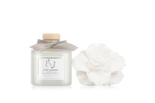 A stylish Marigold Ceramic Flower Diffuser Gift Set - White Jasmine, featuring a frosted glass bottle with a wooden cap and gray ribbon, is elegantly displayed next to a large white flower sculpture. Encased in an elegant box, it makes an ideal gift with a minimalistic design that highlights the floral fragrance theme.