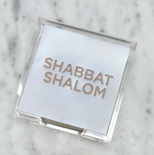 Load image into Gallery viewer, The Cocktail Napkin Acrylic Tray Set - Year Round Entertaining includes a white disposable napkin with &quot;SHABBAT SHALOM&quot; printed in gold letters, all beautifully arranged inside a clear acrylic holder. This stylish setup makes for a thoughtful hostess gift as the holder sits elegantly on a light gray marbled surface.
